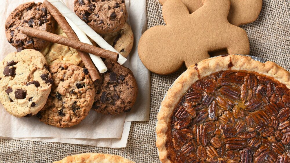 An assortment of baked goods is displayed on a burlap surface. The items include chocolate chip cookies, wafer rolls, gingerbread cookies, and a pecan pie. The cookies are stacked on a napkin, and the pie rests in a foil pan.