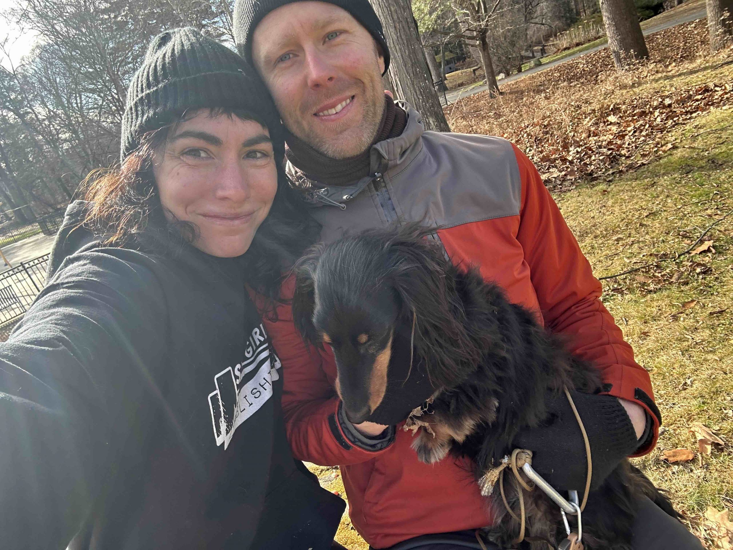 A woman and a man are taking a selfie outdoors while holding a small black dog. They are both wearing beanies, and the man is dressed in a red and gray jacket. The background includes trees and dried leaves on the ground.