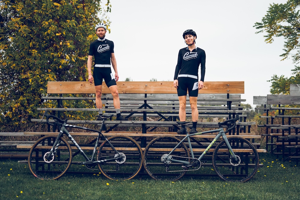 Two cyclists stand on a bench beside their bicycles in a park, wearing matching black and white cycling outfits during their bike training.
