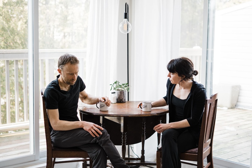 Two people sitting at a round table with mugs in front of them, engaged in a serious conversation about bike coaching in a room with large windows and sheer curtains.