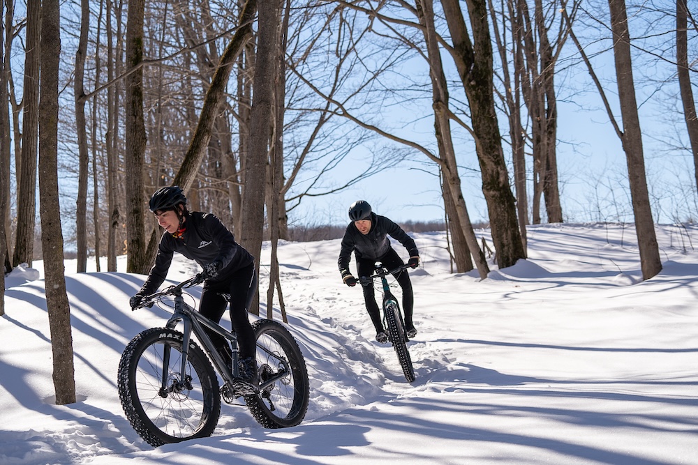 Two cyclists undergoing cycling training on mountain bikes on a snowy forest trail.