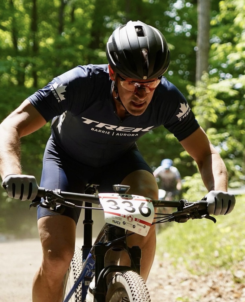 A cyclist wearing a black helmet and a blue kit with the number 336 is intently focusing on bike training while riding a mountain bike on a sunny forest trail.