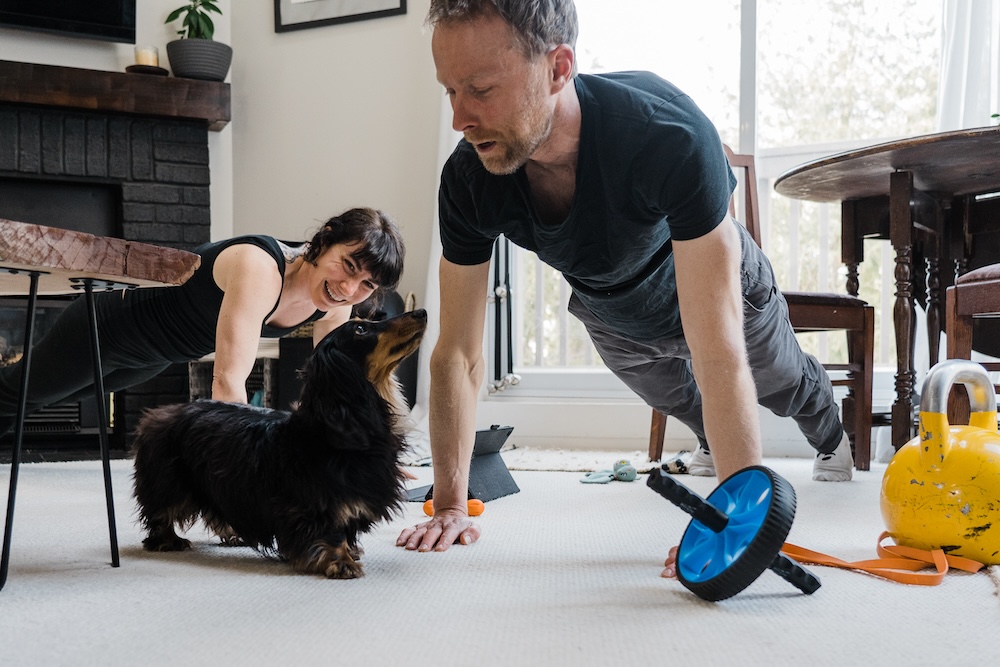 A couple is engaging in a mobility training workout in their living room, with the man doing a push-up while giving a high-five to their dog, as the woman smiles at them, and various exercise