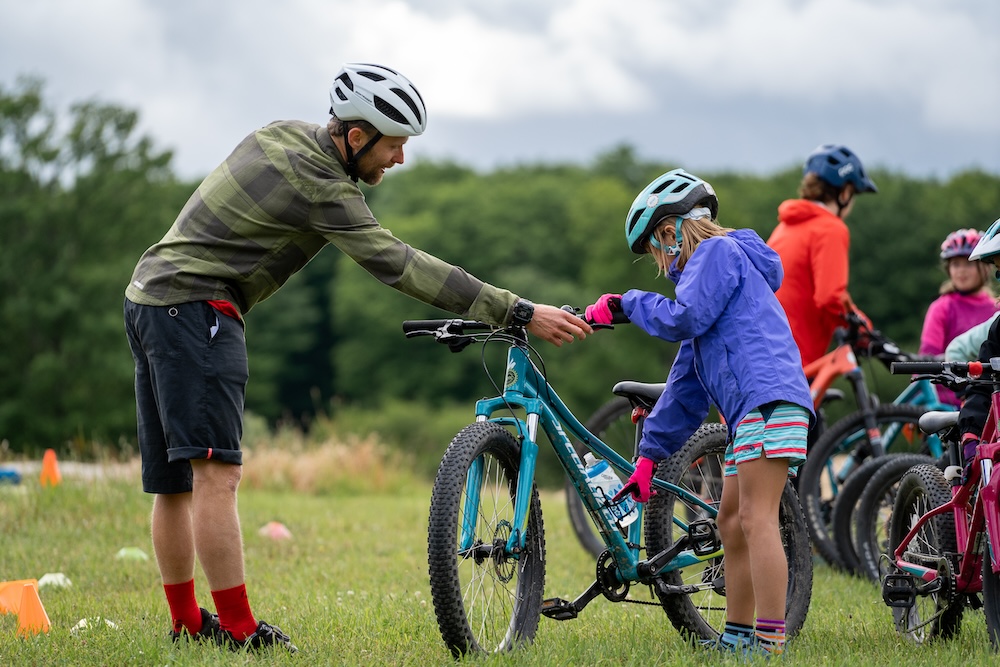 An instructor assists a young girl with her bicycle during a bike training session in a grassy outdoor area.