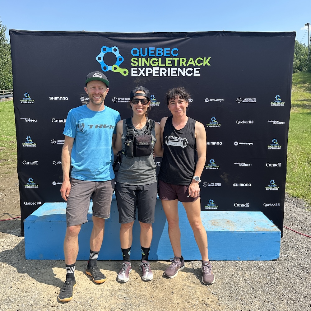 Three people posing together in front of a banner for "Quebec Singletrack Experience," smiling for a photo on a sunny day after their cycling training session.