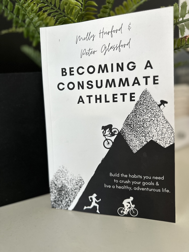 A book titled "becoming a consummate athlete" by molly hurford & peter glassford stands upright on a desk. the cover features a stylized mountain and cyclist graphic.