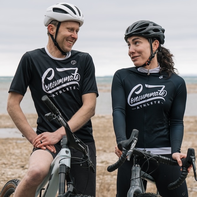 Two cyclists, a man and a woman, wearing black and white cycling gear labeled "consummate athlete," are talking and smiling beside their bikes on a sandy beach.