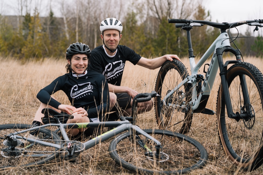 Two cyclists in black and white attire sitting on the ground beside their mountain bikes in a grassy field, smiling at the camera.
