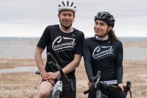 Two cyclists in black and white gear, wearing helmets, stand beside their bicycles on a pebble beach with a cloudy sky and lake in the background.