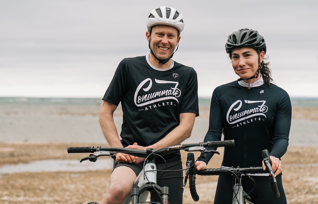 Two cyclists, a man and a woman, smiling and standing on a pebble beach with their bikes. they are wearing helmets and matching black cycling outfits with the word "crankmate" on them.