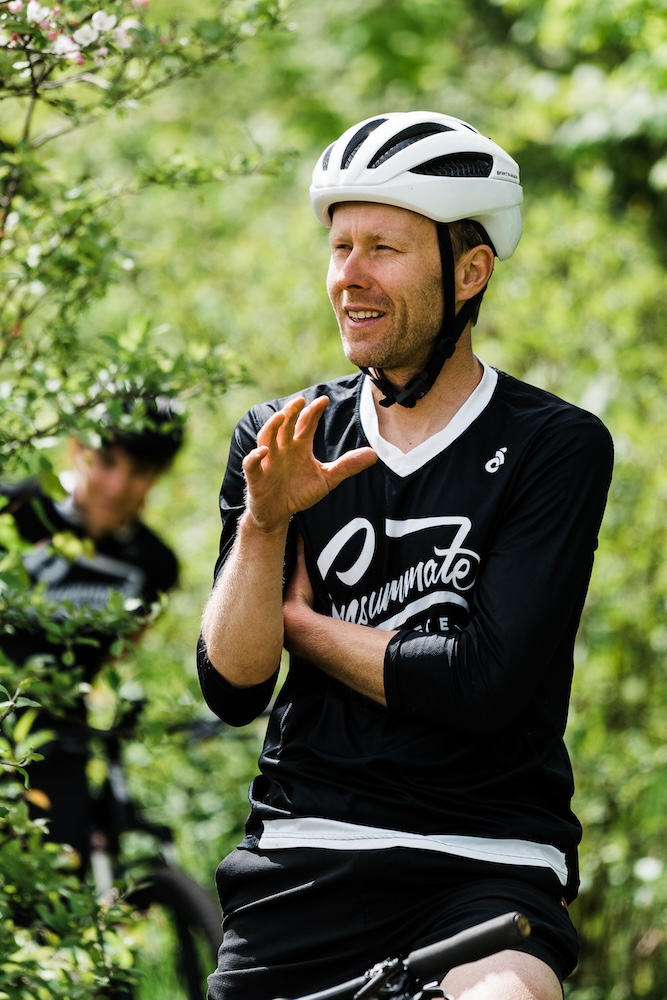 A man in a black cycling jersey and white helmet gesticulates with his hand while smiling, surrounded by lush greenery. another cyclist, out of focus, is seen in the background.