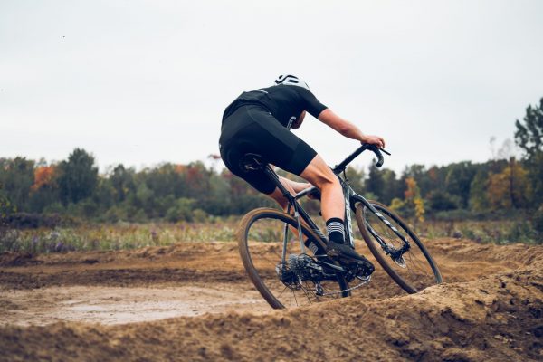 A cyclist in black athletic wear and a helmet rides a gravel bike on a curvy dirt trail, with trees displaying autumn colors in the background.