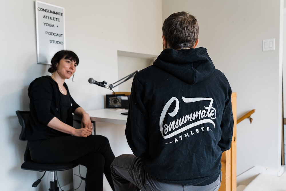A man with his back to the camera, wearing a hoodie labeled "consummate athlete," sits facing a woman who is speaking. they are in a room with a poster that includes the text "athlete, yoga, podcast studio.