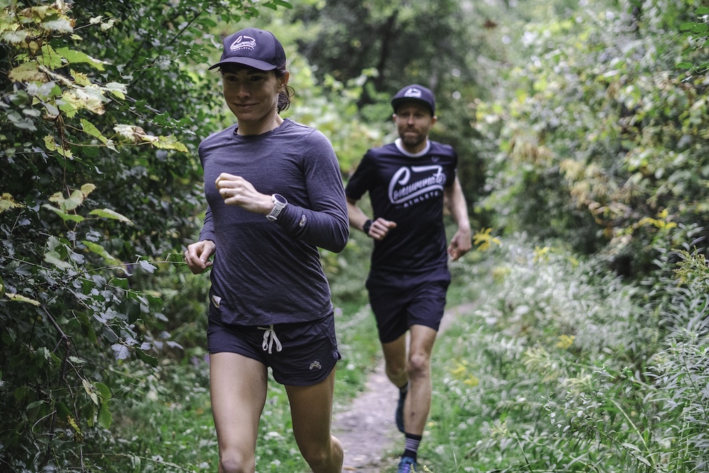 A woman and a man, both in athletic wear, jog on a narrow, leafy trail. the woman is in the foreground and focuses intently ahead, while the man follows behind.
