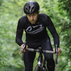 A focused cyclist in black cycling gear riding a road bike on a lush, green forest path. the cyclist is wearing a helmet and looking intently ahead.