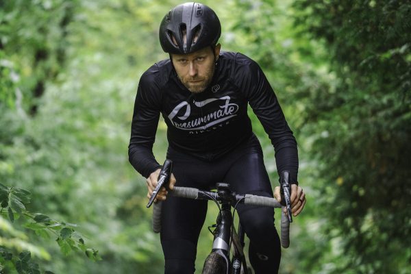 A focused cyclist in black cycling gear riding a road bike on a lush, green forest path. the cyclist is wearing a helmet and looking intently ahead.