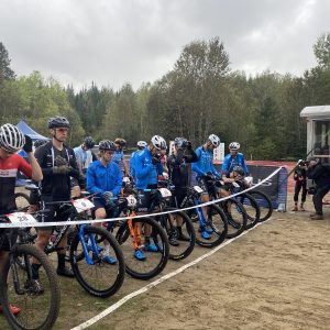 A group of cyclists wearing various cycling gear and helmets is gathered behind a starting line at an outdoor biking event, with a forest in the background.