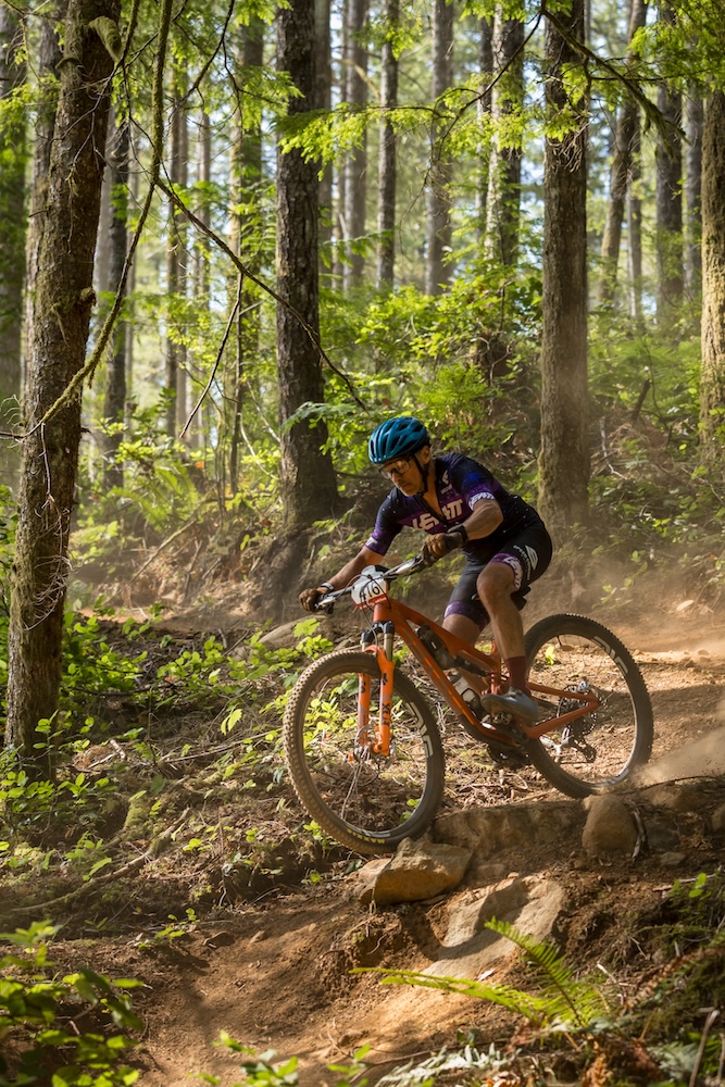 A mountain biker maneuvers a sharp turn on a forest trail, sunlight filtering through the trees, highlighting dust in the air. she is focused and wearing a purple jersey and safety gear.