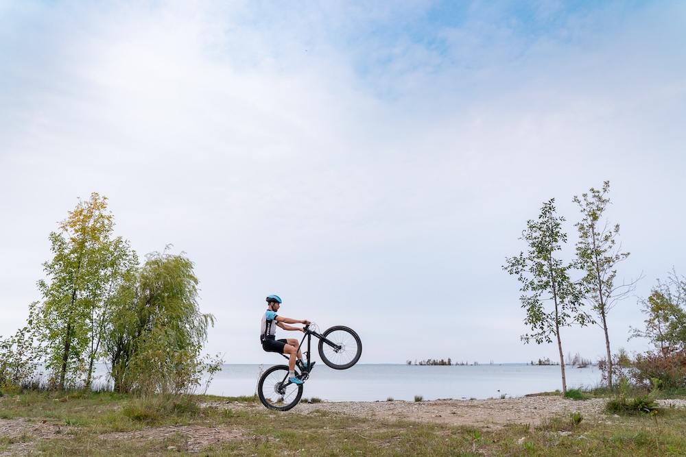 A cyclist performs a wheelie on a dirt path near a lake, surrounded by sparse foliage under a clear sky.