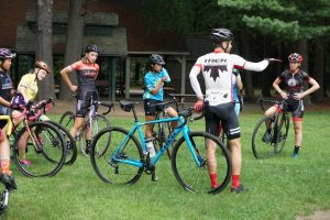 A group of cyclists in various colorful outfits, each standing beside their road bikes, listens attentively to a man pointing and giving instructions in a park setting.
