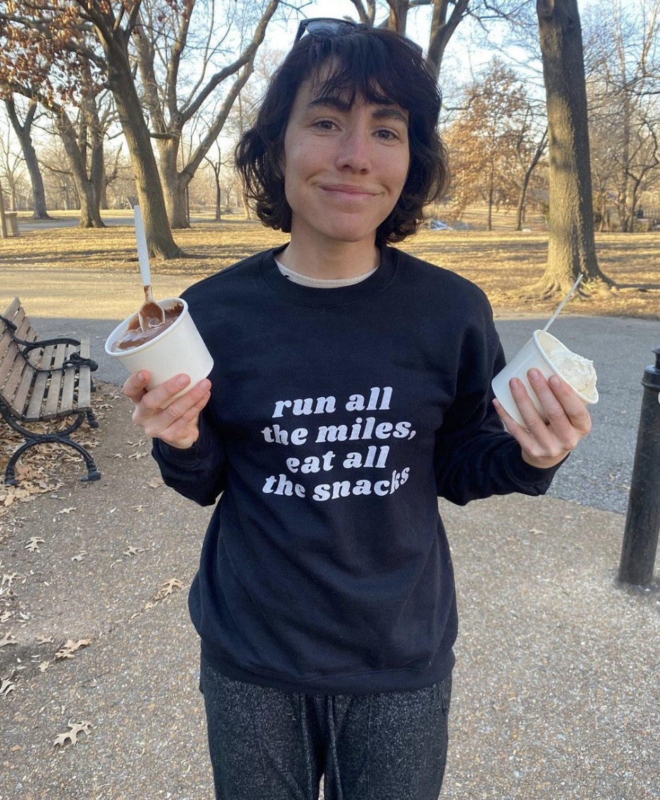 A woman with a joyful expression holding two cups of ice cream in a park. she wears a black sweatshirt with the text "run all the miles, eat all the snacks". trees and a bench are visible in the background.