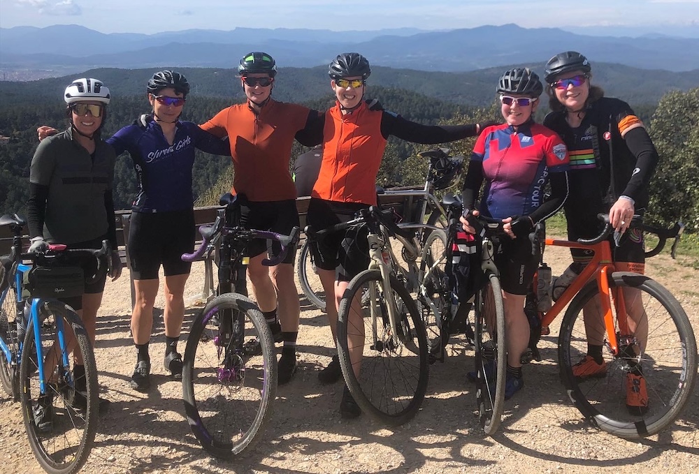 Six cyclists posing with their bikes on a sunny day atop a hill, showing a scenic forested landscape in the background. all are dressed in cycling gear and helmets, smiling at the camera.