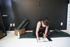 A woman in a black tank top and leggings sits on a yoga mat in a room with dark walls, writing in a notebook. yoga mats and a woven basket are nearby.