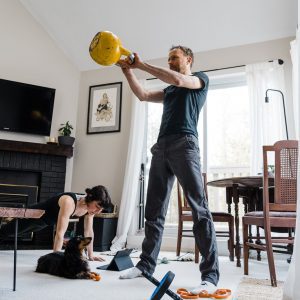 A man lifting a yellow kettlebell in a home living room, while a woman performs a plank exercise beside him. a small dog sits nearby on the floor, and gym equipment is scattered around the room.