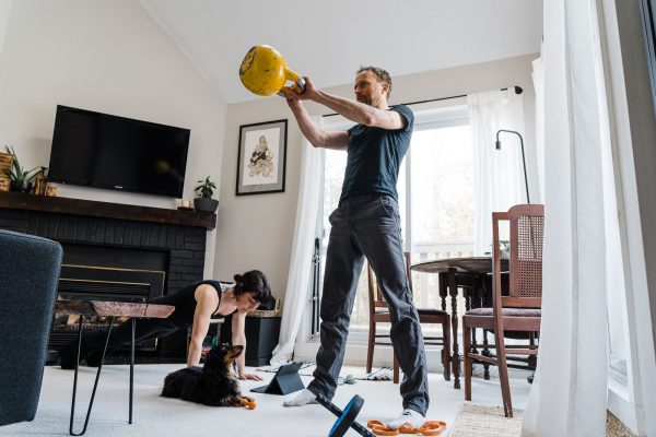 A man lifting a yellow kettlebell in a home living room, while a woman performs a plank exercise beside him. a small dog sits nearby on the floor, and gym equipment is scattered around the room.