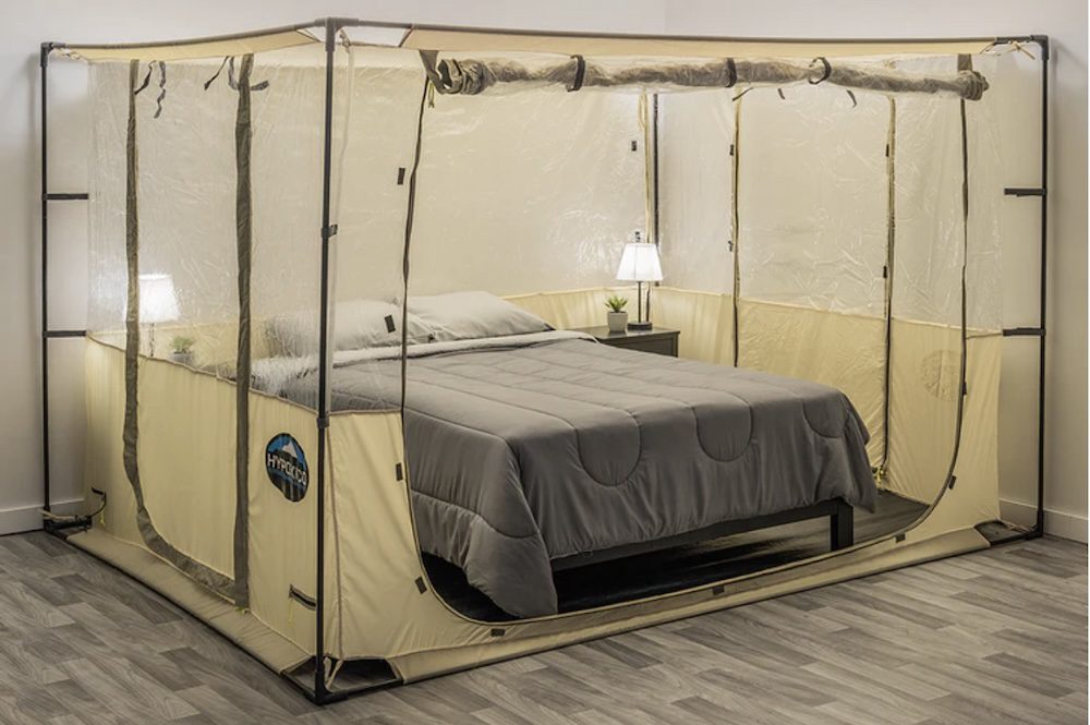 A spacious, beige tent enclosing a bed in a room, featuring clear plastic windows and a bedside table with a lamp, set on a wooden floor.