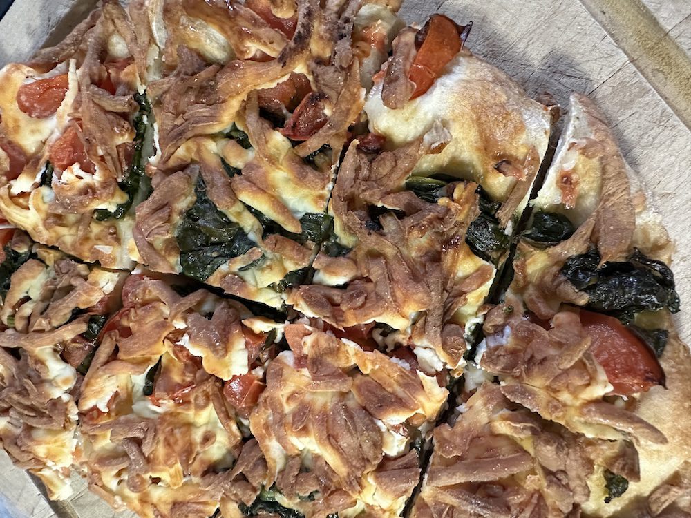 A close-up shot of a homemade spinach and cheese pizza with pieces of caramelized onion on top, served on a wooden board. some slices have been removed, showing the toppings and crust texture.