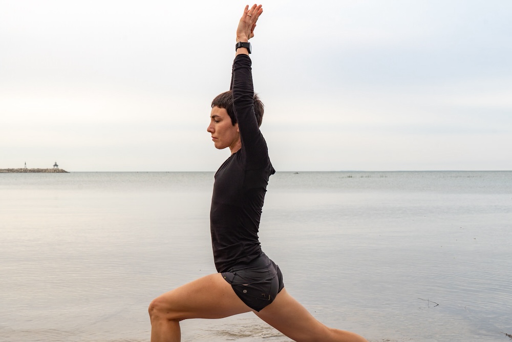 A woman in sportswear performs a yoga pose on a sandy beach with the ocean and a distant lighthouse in the background. she is focused and balanced, with one arm extended upward.