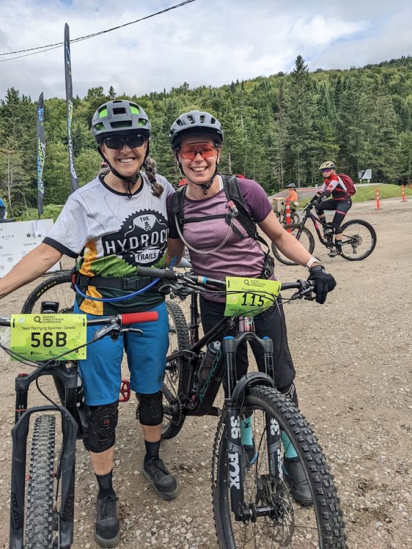 Two cyclists pose with their mountain bikes in an outdoor setting. They wear helmets and biking gear, and both have race bibs on their bikes. Other cyclists and a forested area are visible in the background. It appears to be a sunny day.