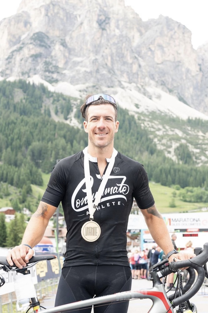 A smiling male athlete wearing a black jersey and a medal stands with a bicycle. mountains and trees are visible in the background. he wears sunglasses on his head.