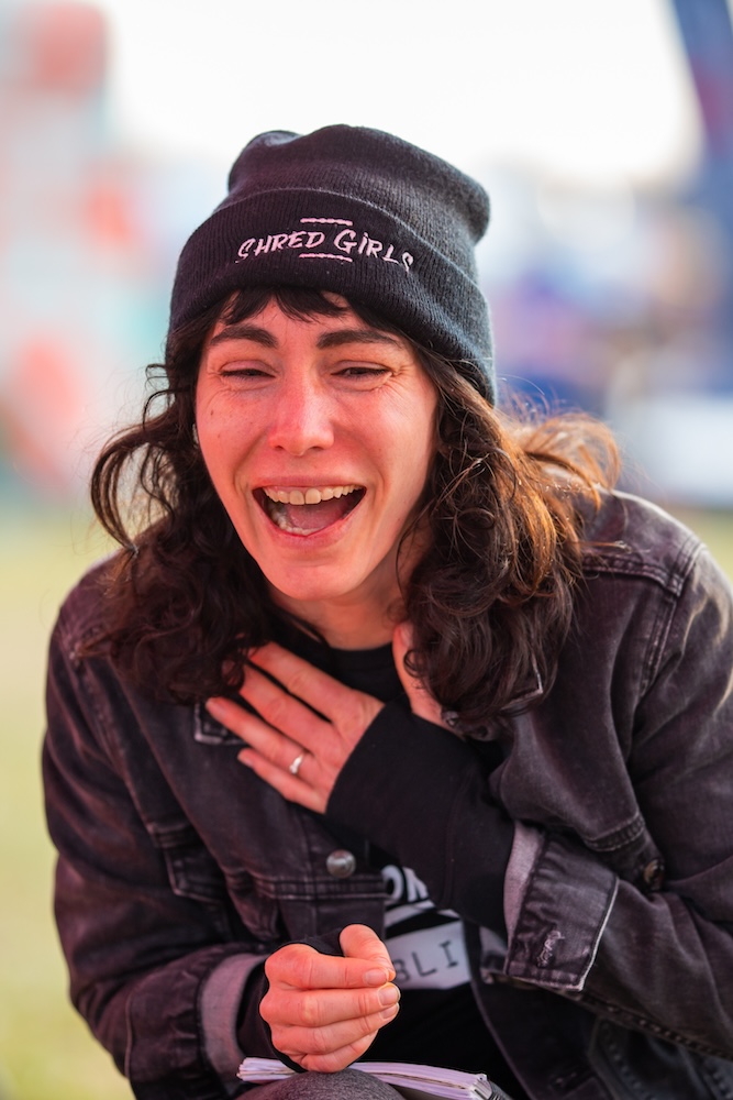 A woman in a denim jacket and a beanie labeled 'cursed girl' laughs heartily, holding her chest, at an outdoor event.