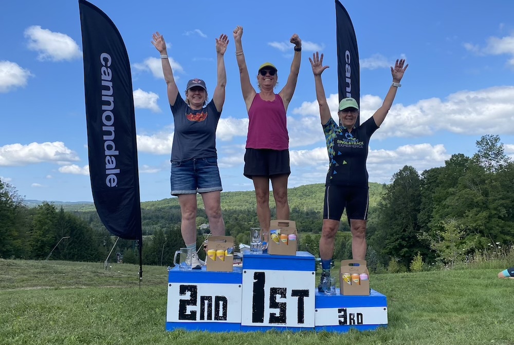 Three women standing on a winners podium outdoors, raising their arms in victory. they are positioned as 1st, 2nd, and 3rd place, surrounded by sponsor banners and a scenic landscape.