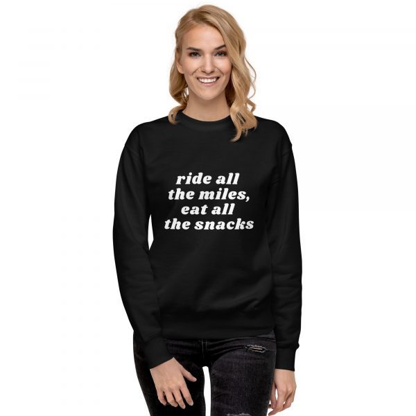 A person with long, blonde hair stands smiling. They are wearing a black sweatshirt with white text that reads, "ride all the miles, eat all the snacks." They are also wearing black jeans with a distressed pattern.