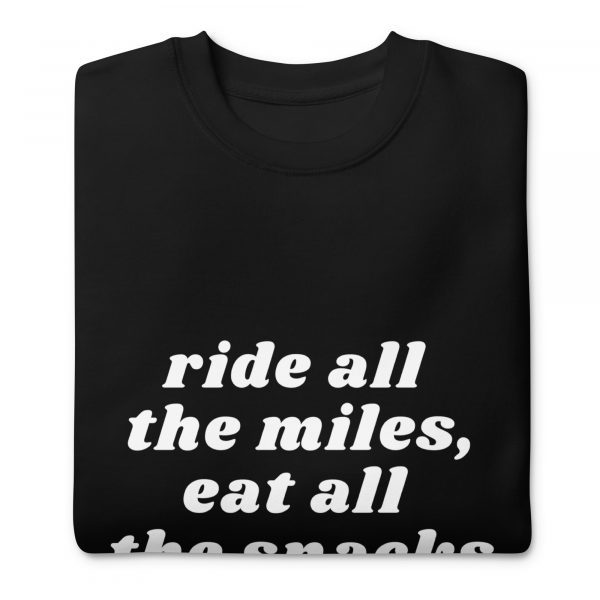 A folded black sweatshirt with a crew neck and white text on the front. The text reads: "ride all the miles, eat all the snacks".