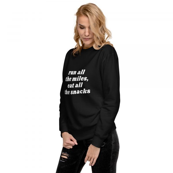 A person with blonde hair is wearing a black sweatshirt that says "run all the miles, eat all the snacks" in white text. The individual is standing against a light-colored background and is looking down with hands loosely clasped in front.