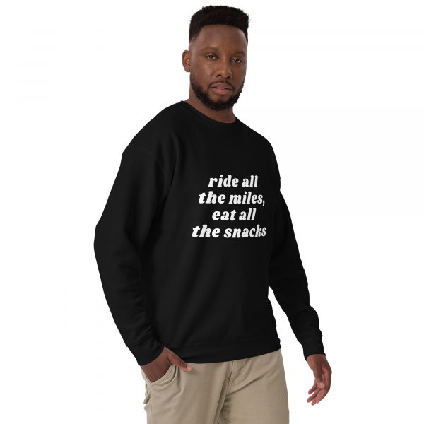 A man with a beard is standing and looking at the camera. He is wearing a black sweatshirt with the text "ride all the miles, eat all the snacks" printed in white on the front. He is also wearing beige pants. The background is white.