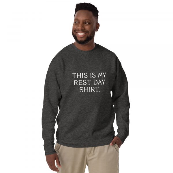 A man with short hair and beard is smiling and wearing a dark gray sweatshirt with the text "THIS IS MY REST DAY SHIRT" printed on the front. He is also wearing beige pants and has one hand in his pocket. The background is plain and white.